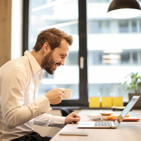 A male executive smiles while holding a cup and reviewing a briefing on his laptop.