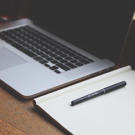 A wooden table with with a laptop, notebooks, and a pen.