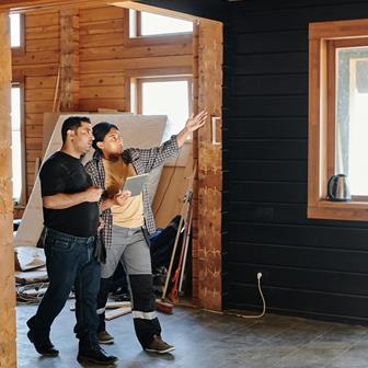 A security expert and a project manager stand in a partially renovated wooden room, discussing plans while one gestures toward a window.