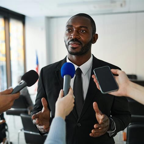 An executive in a suit speaks to reporters holding microphones and a phone in a professional setting.