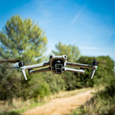 A drone hovers mid-air above a dirt path in a natural outdoor setting with trees in the background.